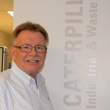 Head shot of Tom Griffith grey hair, square glasses standing beside a verticle Caterpillar tradeshow white banner